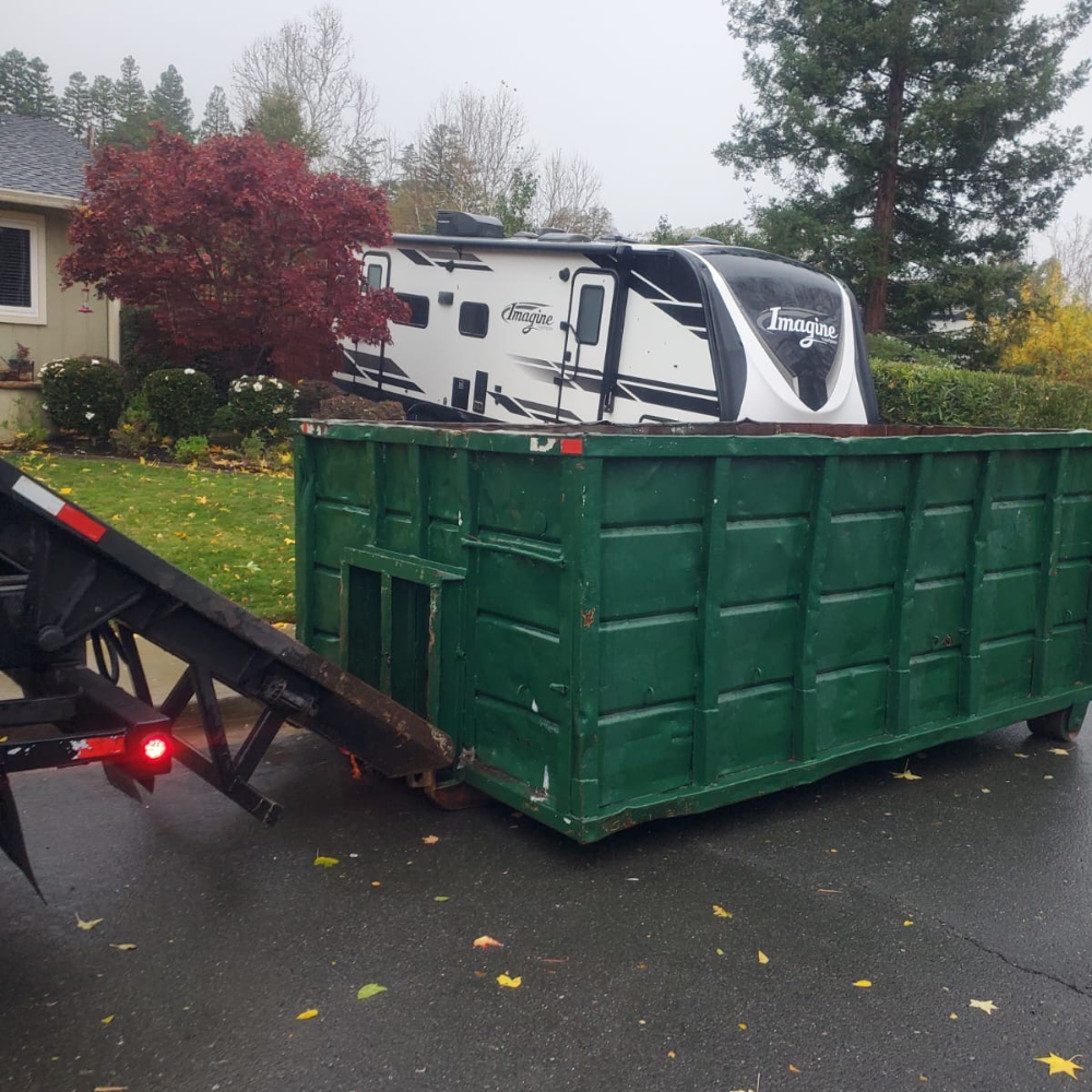Green dumpster being lowered next to an RV in a fall driveway